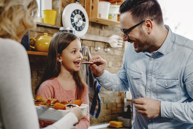 Familia feliz probando comida en la cocina El padre está alimentando a la niña