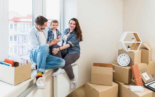 Familia feliz sentada en el alféizar de la ventana con cajas de cartón en movimiento en su nuevo hogar