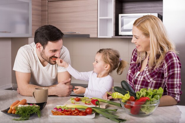 Familia feliz con su pequeña hija haciendo una ensalada fresca con verduras en la cocina