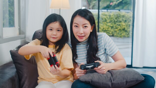 La familia y la hija asiáticas jovenes juegan a juegos en casa. Madre coreana con niña con joystick divertido momento feliz juntos en el sofá en la sala de estar en casa. Madre divertida y niño encantador se divierten