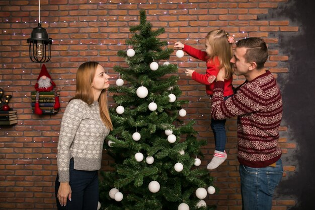 Familia con hija pequeña colgando juguetes en un árbol de Navidad