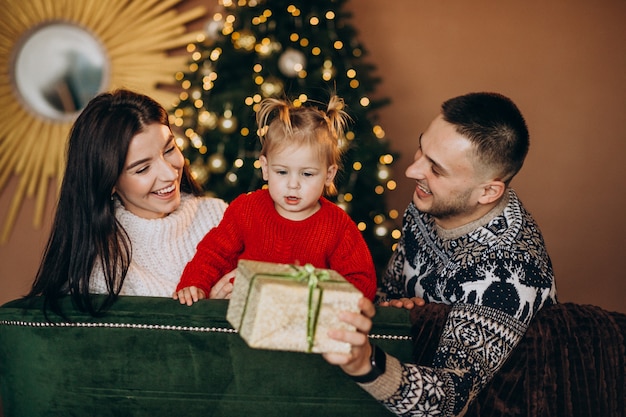 Familia con hija pequeña sentada junto a árbol de Navidad y desempacando caja de regalo