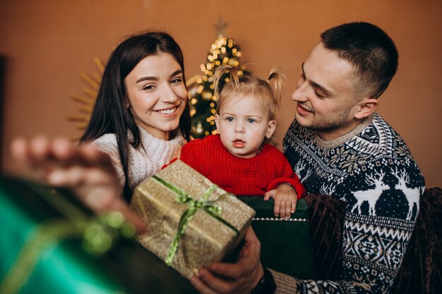 Familia con hija pequeña sentada junto a árbol de Navidad y desempacando caja de regalo