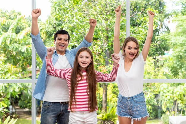 La familia, incluido el padre, la madre y la hija, de pie y abrazándose frente a la ventana de vidrio en casa, levanta la mano con alegría