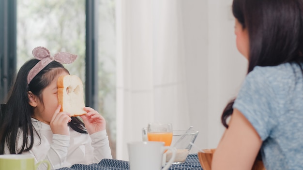 Familia japonesa asiática desayuna en casa. La hija asiática escoge y juega la sonrisa de risa del pan con los padres mientras come cereal de cereales y leche en un tazón sobre la mesa en la cocina moderna en la mañana.