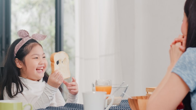 Familia japonesa asiática desayuna en casa. La hija asiática escoge y juega la sonrisa de risa del pan con los padres mientras come cereal de cereales y leche en un tazón sobre la mesa en la cocina moderna en la mañana.