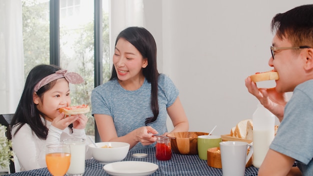 Familia japonesa asiática desayuna en casa. La mamá feliz asiática que hace la mermelada de fresa en el pan para la hija come cereal y leche de las avenas en cuenco en la tabla en la cocina en la mañana.