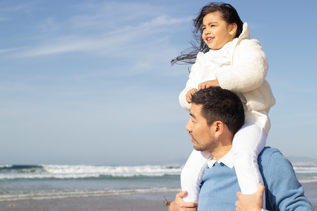 Familia japonesa contenta caminando por la playa. Hija cabalgando sobre la espalda de los padres, mirando hacia adelante. Ocio, vacaciones, concepto de crianza