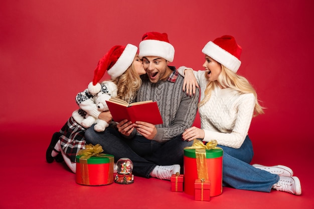 Familia joven alegre conmocionada que lleva el libro de lectura de los sombreros de la Navidad.