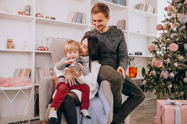 Familia joven con pequeño hijo sentado junto al árbol de Navidad desempacando regalos