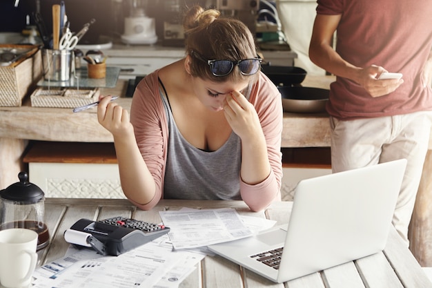 Familia joven que enfrenta problemas de deuda. Mujer estresada sosteniendo su cabeza con frustración, haciendo cuentas bancarias y haciendo los cálculos necesarios usando una computadora portátil y una calculadora