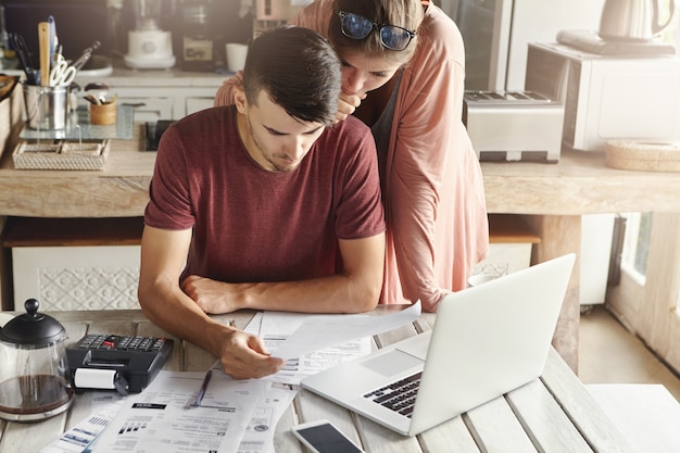 Foto gratuita familia joven que maneja el presupuesto, revisa sus cuentas bancarias usando una computadora portátil genérica y una calculadora en la cocina. marido y mujer haciendo trámites juntos, pagando impuestos en línea en una computadora portátil