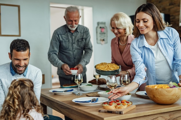 Familia multigeneracional almorzando juntos en casa El foco está en la mujer sonriente que lleva comida y vino a la mesa