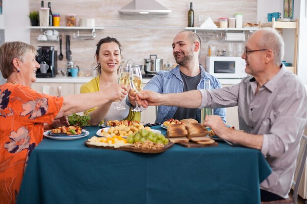 Familia multigeneracional brindando con vino blanco durante el almuerzo. Sabrosas patatas sazonadas. Padres mayores. Uvas deliciosas.