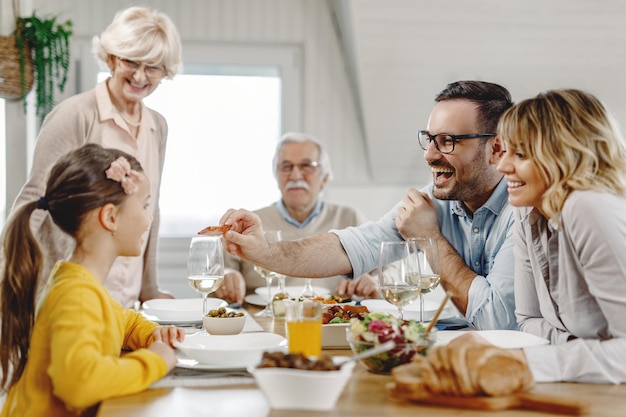 Familia multigeneracional divirtiéndose durante el almuerzo mientras el padre alimenta a su niña en la mesa del comedor