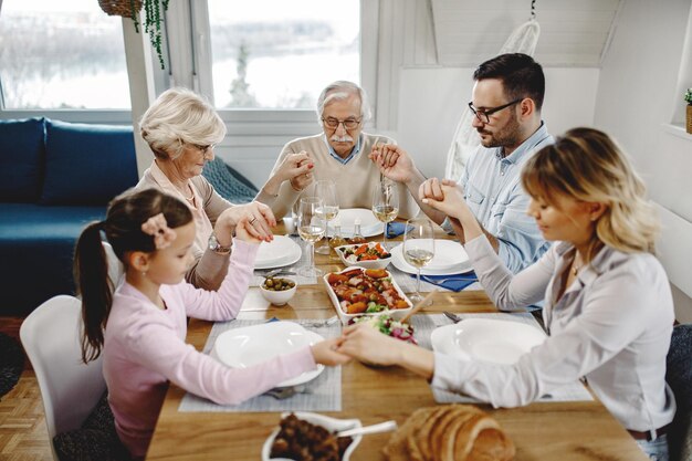 Familia multigeneracional sentada en la mesa del comedor y dando gracias mientras se toma de la mano