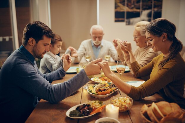 Familia multigeneracional tomados de la mano y rezando durante el almuerzo en el comedor