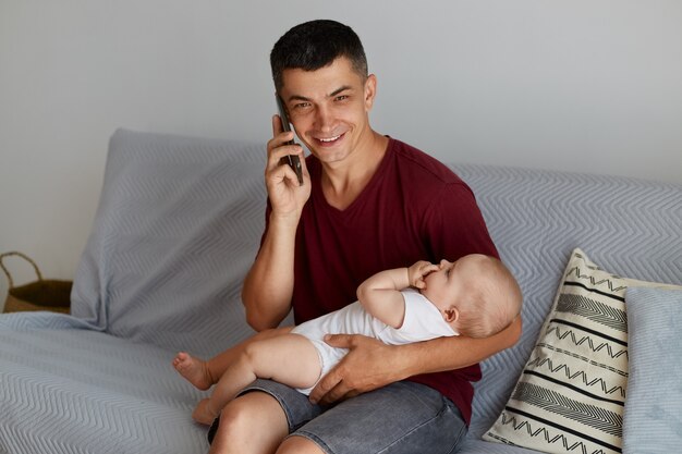 Familia, paternidad e infancia. Retrato de padre sonriente feliz con niño o niña llamando por teléfono inteligente en casa mientras está sentado en el sofá en la sala de estar.