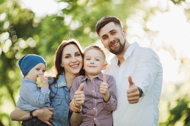 Familia positiva y elegante caminando en el parque junto con niños posando y gesticulando súper con los dedos en la cámara Madre y padre abrazando a sus dos hijos Concepto de aspecto familiar