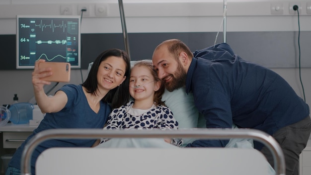 Familia de positividad tomando selfie con smartphone moderno durante la consulta de enfermedad en la sala del hospital. Paciente del niño pequeño descansando en la cama recuperándose después de la terapia de la enfermedad