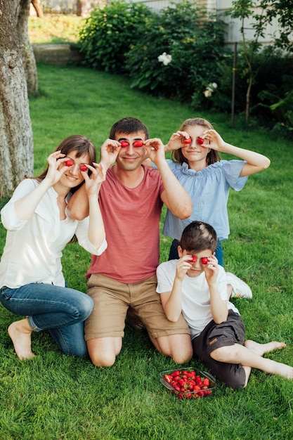 Familia sonriente cubriendo sus ojos con fresas frescas durante un picnic