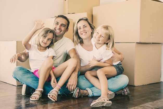 Foto gratuita familia sonriente con niños sentados en el piso cerca de cajas de cartón y relajarse. chica rubia en las piernas del padre agitando