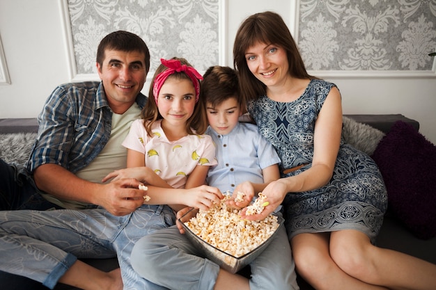 Foto gratuita familia sonriente que sostiene las palomitas y que mira la cámara mientras que se sienta en el sofá