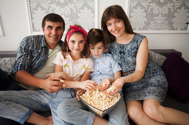 Familia sonriente que sostiene las palomitas y que mira la cámara mientras que se sienta en el sofá