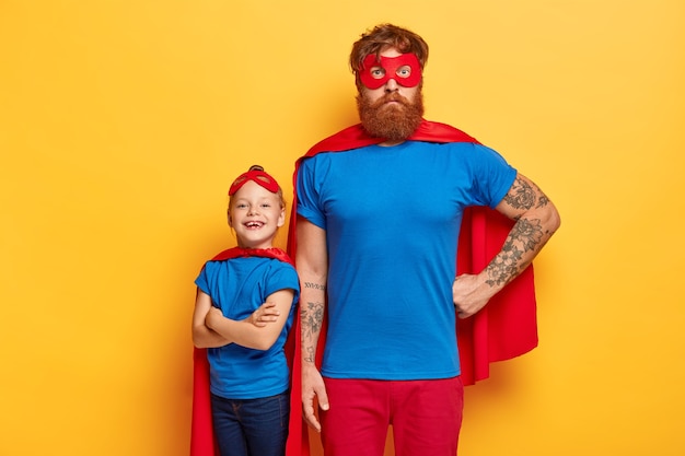 Familia de superhéroes. Papá poderoso mantiene una mano en la cintura, el niño pequeño con los brazos cruzados retrocede