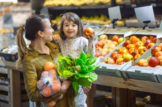 Familia en el supermercado. Hermosa joven mamá y su pequeña hija sonriendo y comprando comida. El concepto de alimentación saludable. Cosecha