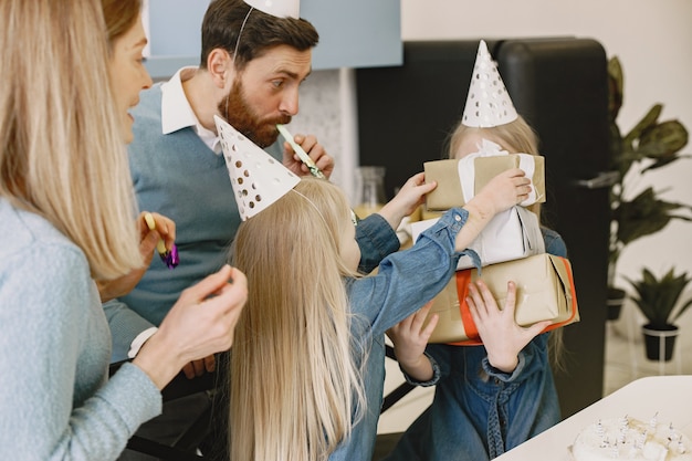La familia y sus dos hijas celebran su cumpleaños en la cocina. La gente usa un sombrero de fiesta. Chica guarda cajas con regalos.