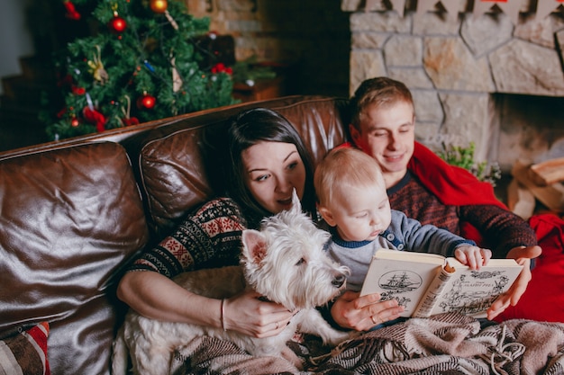 Familia tumbada en un sofá con una manta mientras leen un libro en tiempos de navidad