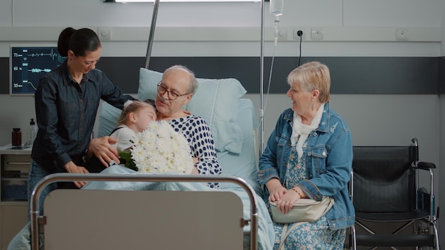 Familia visitando a un paciente mayor y trayendo flores en la cama de la sala del hospital. madre e hijo abrazando a un anciano en una visita familiar a la clínica, disfrutando de la recuperación con su hija y su niña. visitantes