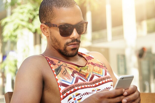 Famoso blogger africano de moda en tonos que se esconde del calor del verano en el parque usando un teléfono inteligente para compartir su nueva publicación a través de las redes sociales, luciendo serio y concentrado. Hombre negro enviando mensajes de texto al aire libre