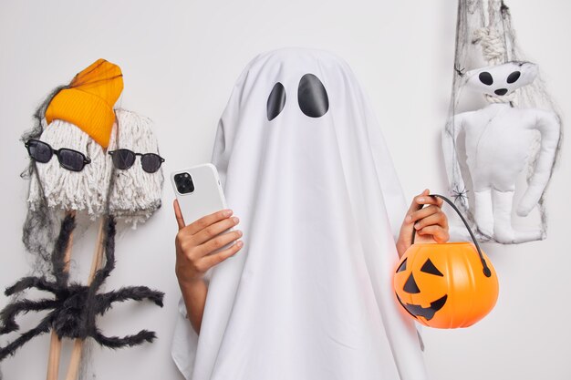 Fantasma femenino irreconocible sostiene un teléfono móvil moderno y una calabaza tallada se prepara para las búsquedas de celebración de Halloween en las ideas de Internet para decorar la habitación antes de las poses de la fiesta cerca de los juguetes de miedo en el interior.