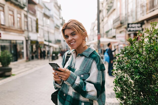 Fantástica mujer bonita con cabello rubio con camisa brillante usando un teléfono inteligente mientras camina por la calle en Europa Chica viajando por Europa en otoño