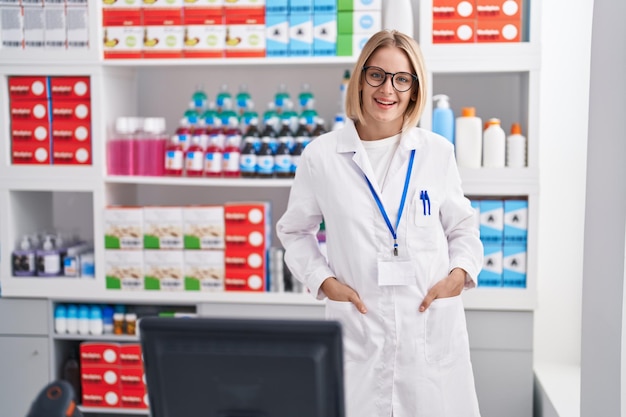 Farmacéutico joven mujer rubia sonriendo seguro de pie en la farmacia