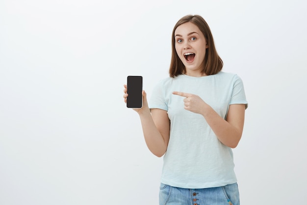 Fascinada y atractiva joven asistente de tienda en camisa casual que muestra la pantalla del teléfono inteligente y apunta al dispositivo hablando sobre nuevas características y diseño fresco, sonriendo posando sobre una pared gris