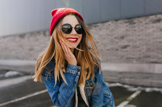 Fascinante chica blanca tocándose la cara y riendo en el fondo urbano Mujer rubia refinada con elegante sombrero rojo pasando tiempo al aire libre en el fin de semana de primavera