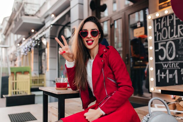 Fascinante chica europea en traje rojo casual divirtiéndose en el café