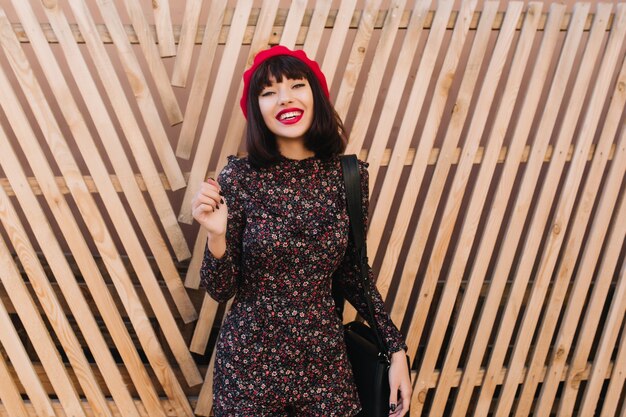 Fascinante chica morena en boina roja de moda y vestido de estilo retro con estampado floral de pie frente a la valla de madera. Adorable joven con corte de pelo corto en traje francés posando al aire libre