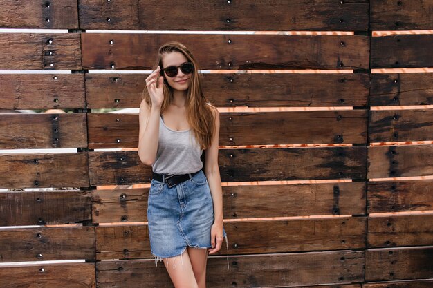 Fascinante chica pálida en gafas de sol de pie sobre una pared de madera. Modelo de mujer blanca alegre en falda de mezclilla relajante.