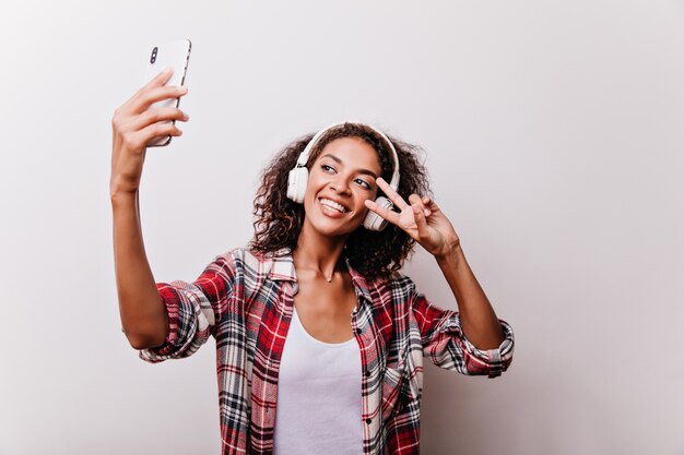 Fascinante chica de pelo negro posando con el signo de la paz para selfie. Mujer joven atractiva en auriculares blancos con smartphone.