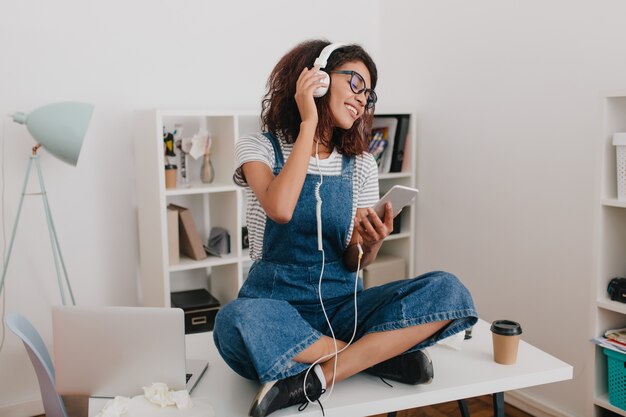 Fascinante chica rizada divirtiéndose en el lugar de trabajo sosteniendo el teléfono inteligente en las manos