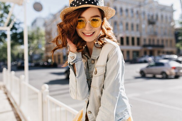 Fascinante joven jengibre en ropa casual posando en la calle. Tiro al aire libre de niña alegre con peinado ondulado que expresa felicidad en el fin de semana de verano.