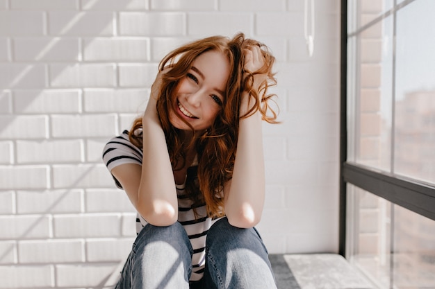 Fascinante modelo caucásico joven posando en casa. Magnífica pelirroja jugando con su cabello y riendo.