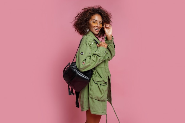 Fascinante modelo de raza mixta en chaqueta verde de moda posando sobre fondo rosa. Gafas de sol amarillas, mochila negra.