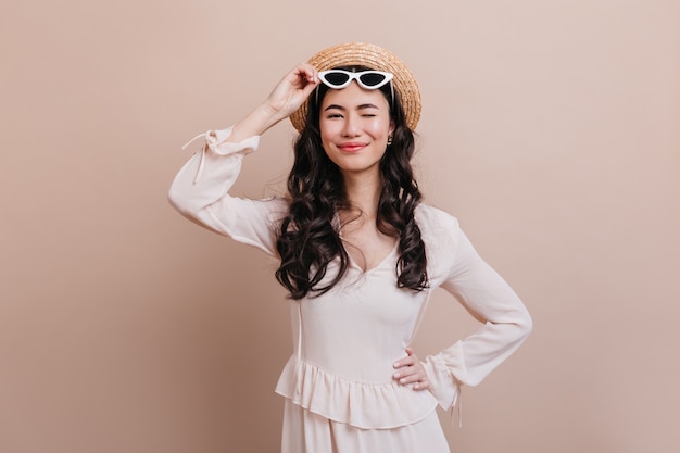 Fascinante mujer asiática sonriendo sobre fondo beige. Vista frontal de la divertida mujer japonesa con sombrero de paja y gafas de sol.