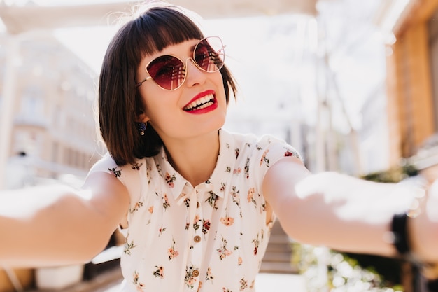 Fascinante mujer caucásica con cabello castaño oscuro haciendo selfie en mañana soleada. Retrato al aire libre de una chica romántica en blusa de moda.