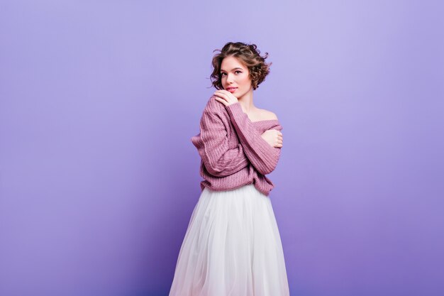 Fascinante mujer europea en camisa de punto suave posando con interés. señorita refinada con peinado elegante aislado en la pared púrpura.